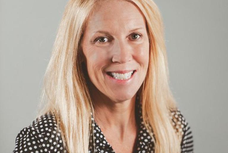 The image shows a woman with long, straight blonde hair wearing a black blouse with white polka dots. 她脸上带着灿烂的笑容，显得很友好 and is facing slightly to her right. The background is a plain light gray. 她皮肤白皙，举止平易近人.