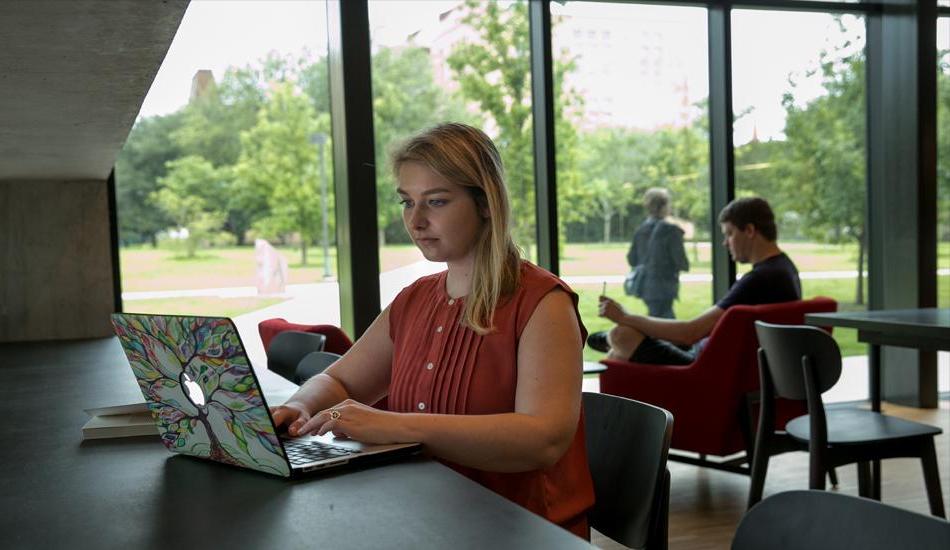 The image shows a young woman with blonde hair, wearing a sleeveless rust-colored blouse, working on a laptop in a modern, spacious room with large windows. Her laptop has a colorful tree design on the cover. She is seated at a long table, focused on her work. In the background, a man is seated in a red chair reading, while another person walks outside in a lush green park. 设置似乎是一个安静，光线充足的书房或休息区，可以看到户外景色.